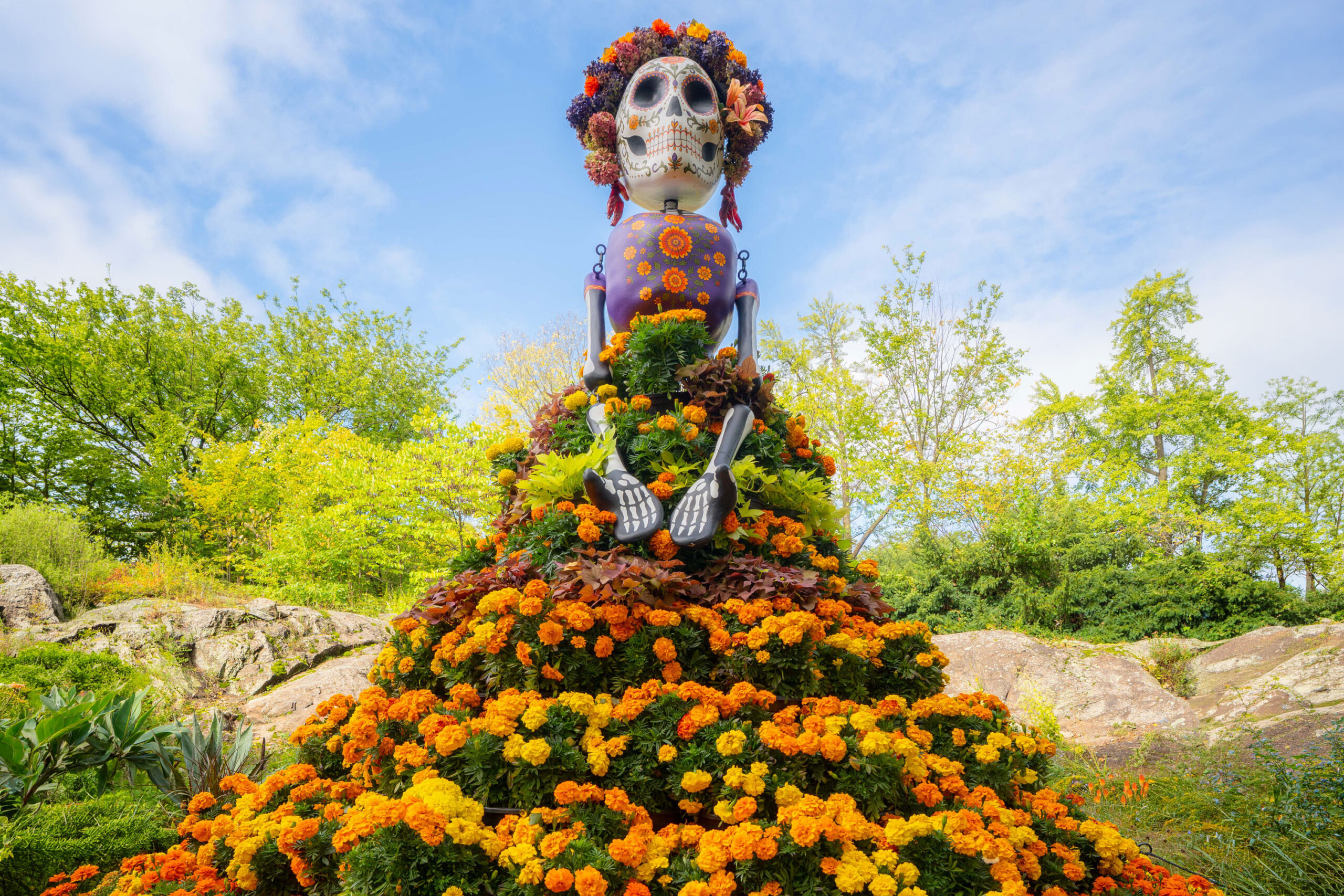 A tall, stylized sculpture of a skeleton, wearing a dress of orange and purple flowers and a flower crown, under a sunny blue sky