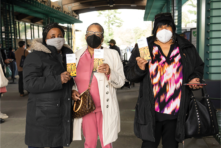 Three visitors pose with their printed exhibition pamphlets from KUSAMA: Cosmic Nature, with other visitors and Garden ticket windows visible behind 