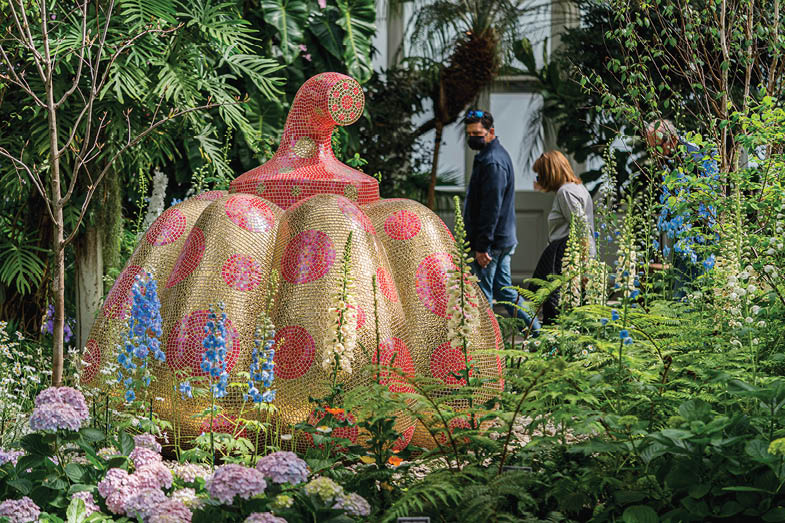 Several visitors stop to look at a glittering statue of a pumpkin in gold with red polka-dots, surrounded by blooming purple hydrangeas and green foliage in the Enid A  Haupt Conservatory 