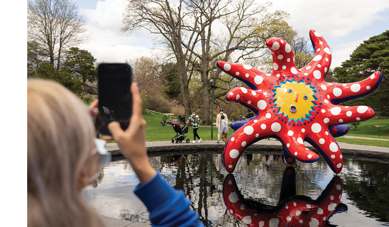 A visitor in the foreground takes a photo of a vividly colored statue featuring a face of surprise, with many red and white polka-dotted arms radiating from it like sunbeams 