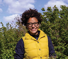 Headshot of Makalé Cullen wearing a black shirt and yellow vest in front of a hedgerow