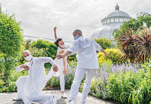 Dancers in flowing white outfits perform choreography in the summer display of the Perennial Garden, surrounded by leafy green foliage with the Enid A  Haupt Conservatory visible in the background 
