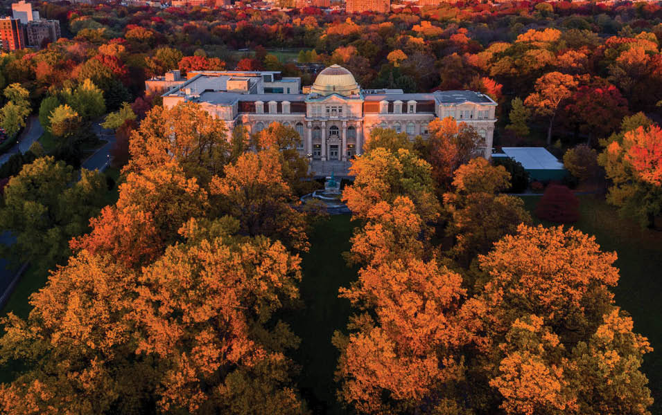 A dramatic photo of the LuEsther T  Mertz Library taken from above, surrounded by the orange, yellow, and red foliage of fall trees as the sun sets in the Bronx