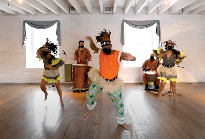 Dancers perform a choreographed dance to the beat of nearby drummers in a brightly lit room